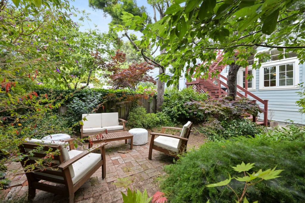 The backyard of 138 27th St, a Noe Valley condo sold by Vivre Real Estate, with a brick paver patio and wood chairs, surrounded by lush green landscaping.