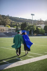 View of Two High School Graduates with Cap and Gowns On While Walking on Football Field