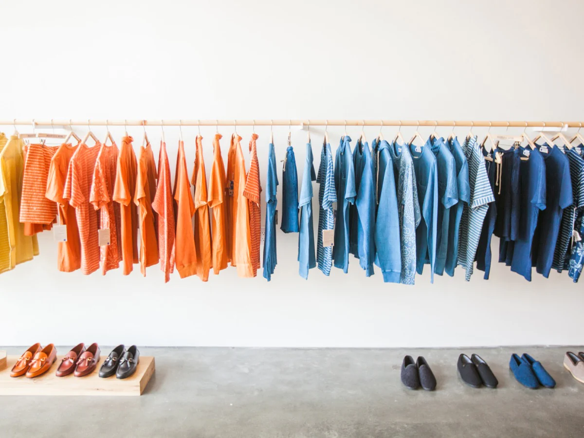Wood rack full of yellow, orange, and blue cotton apparel including t-shirts and shorts, plus an array of leatherlike dress shoes and slip on suede shoes.