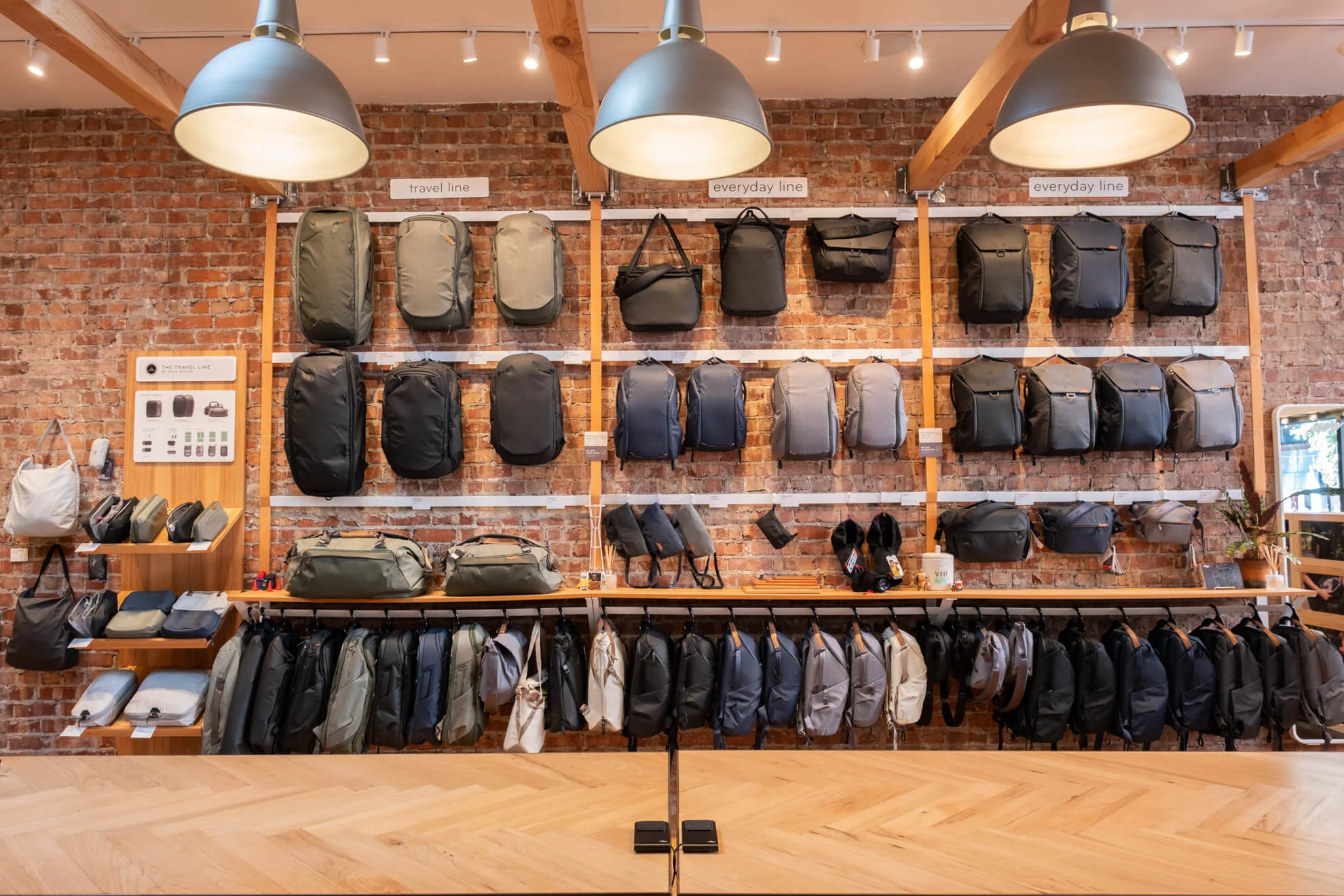 Handmade quality backpacks and bags in neutral colors including black, gray, tan, and khaki aligning a brick wall inside Peak Design San Francisco