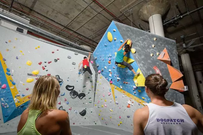 Man in Red Tank Top and Woman in Teal Tank Top Climbing a Rock Climbing Wall while Two Women Watch, one wearing a Dogpatch Boulders White Tank