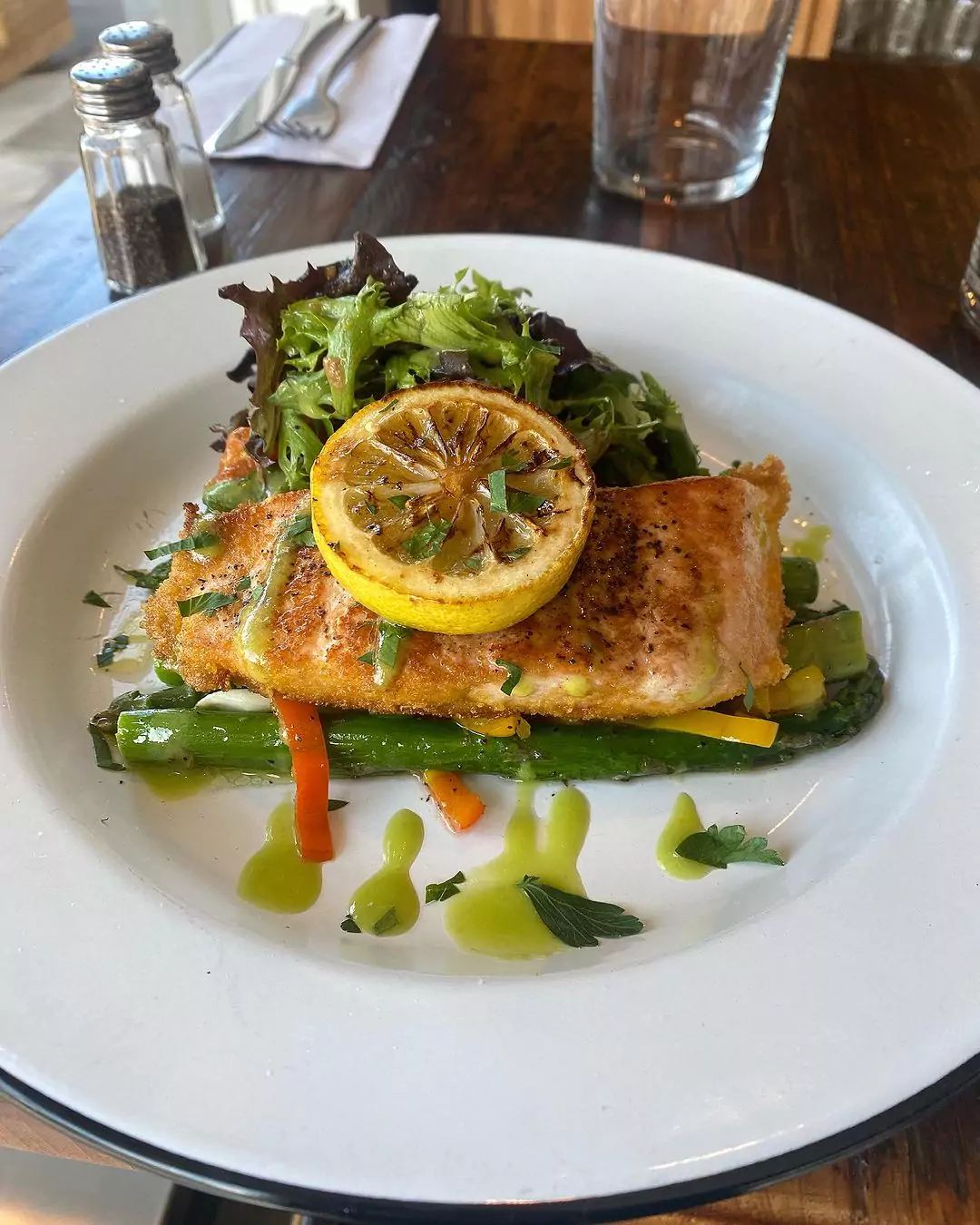 Plate of roasted salmon topped with grilled lemon half and served atop asparagus with a green sauce and side mixed green salad