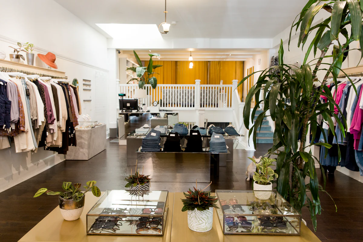 Interior view of shop filled with clothing racks full of items for sale, sunglass cases, stacks of jeans, and numerous plants at Two Birds San Francisco