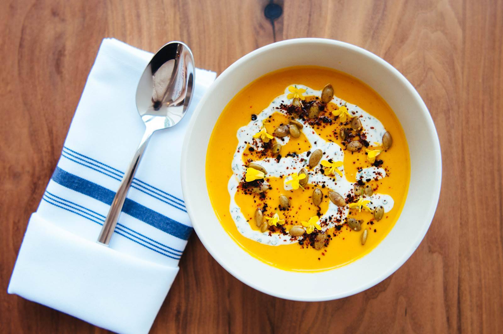 Bowl of Butternut Squash Soup topped with Pumpkin seeds, cayenne dusting, edible flowers, and a white cream sauce with folded white and blue stripe napkin with single spoon beside bowl