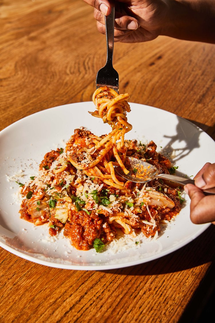 Close up of diner’s hands digging into a plate of spaghetti with red meat sauce and topped with herbs and grated parmesan cheese from Nopa