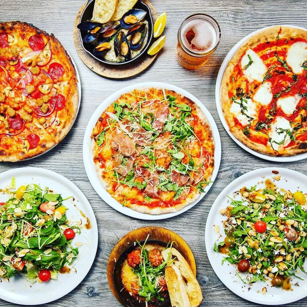 Wood Table topped with multiple red sauce pizza pies including a margherita and arugula and prosciutto pie, alongside two large family-style salads, and meatballs in red sauce with bread for dipping