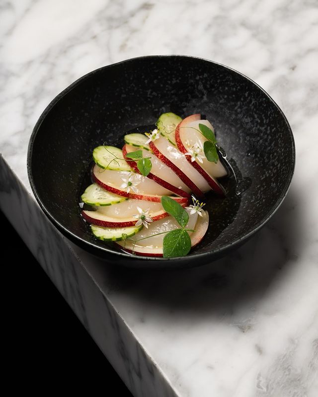 Black bowl filled with sliced hokkaido scallop, sliced nectarine and cucumber, topped with microgreens and edible white flowers at fine dining establishment Commis