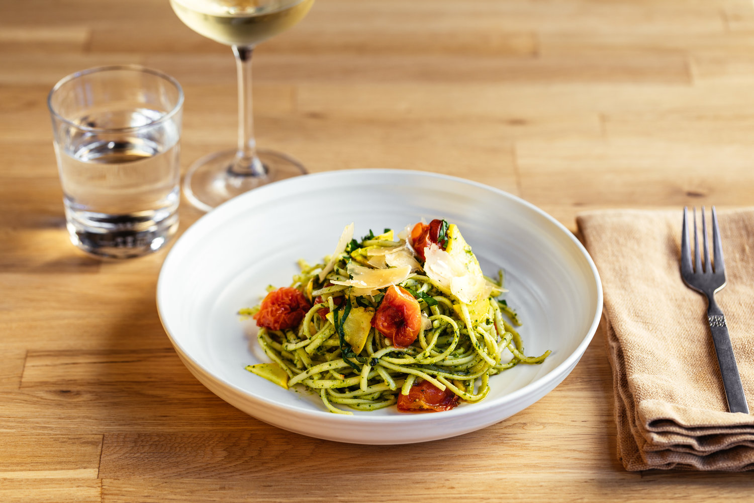 Dish of spaghetti with green sauce, tomatoes, and grated parmesan cheese, with a sliver of a wine glass filled with white wine in the background