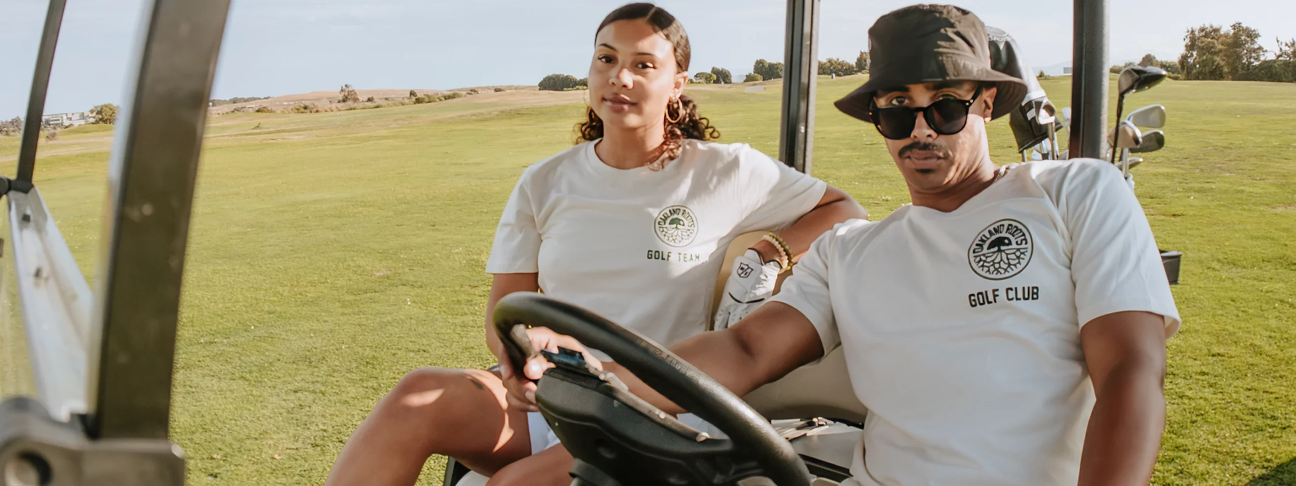 Male and Female sitting on golf cart wearing light colored tee shirts with “Oakland Roots” printed on them