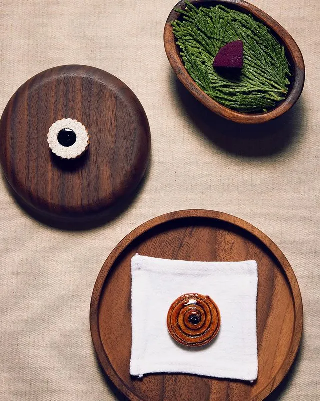 A trio of bite-sized sweet treats served on dark wood plates including a glazed rolled bun, a powdered sugar-coated shortbread cookie filled with purple-colored jam, and a ruby jelly slice