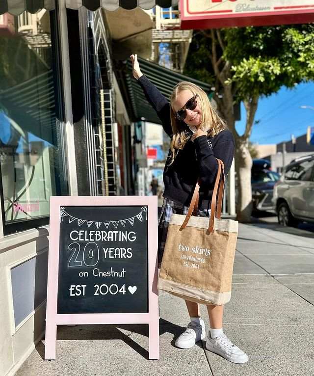 Photo of young girl in blue pullover, checkered blue skirt, white sneakers, and large brown tortoise sunglasses and holding a “Two Skirts” canvas tote bag, while standing on sidewalk beside a chalkboard sign that reads “Celebrating 20 Years on Chestnut, Est. 2004”