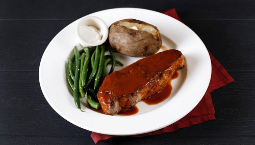 Plate of steak with brown sauce, baked potato with a side of sour cream in round white dish and green beans topped with salt from The Brazen Head