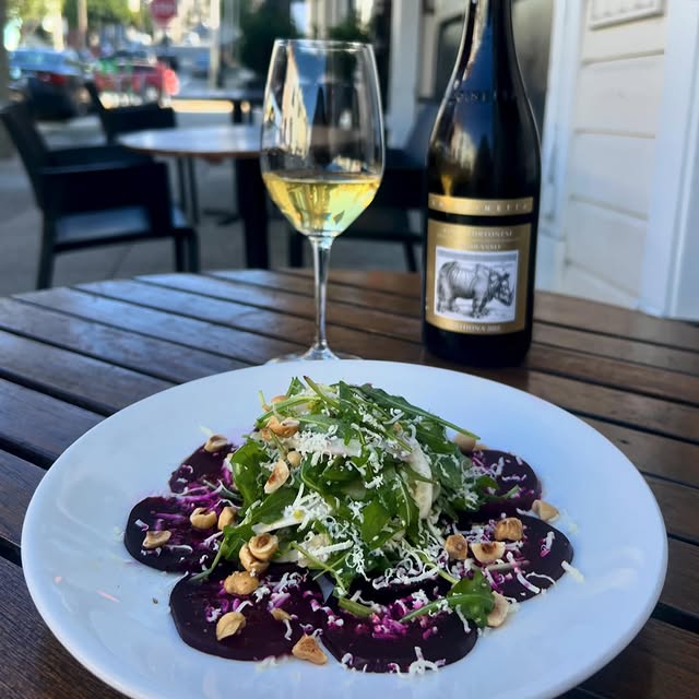 Plate of thinly sliced beet carpaccio topped with chopped hazelnuts, grated parmesan cheese, and arugula, with a bottle and glass of white wine set on an outdoor sidewalk table at Terzo in Cow Hollow