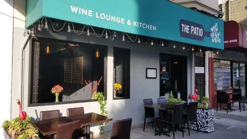 View from sidewalk of small restaurant space with two four-person outdoor tables and a green awning over the windows which states “The Patio” in a wine bottle graphic and also “Wine Lounge & Kitchen”