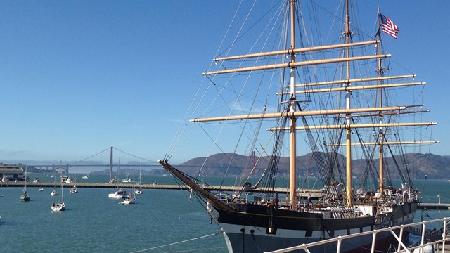 View of a historic ship topped with an American flag at Hyde Street Pier, with views of the bay and Aquatic Cove in the background