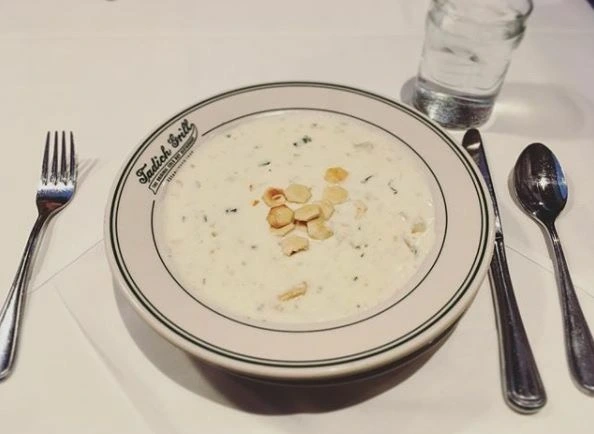 Bowl of Boston Clam Chowder topped with oyster crackers and served in a white bowl that says “Tadich Grill”, atop a white tablecloth with flatware and a glass o