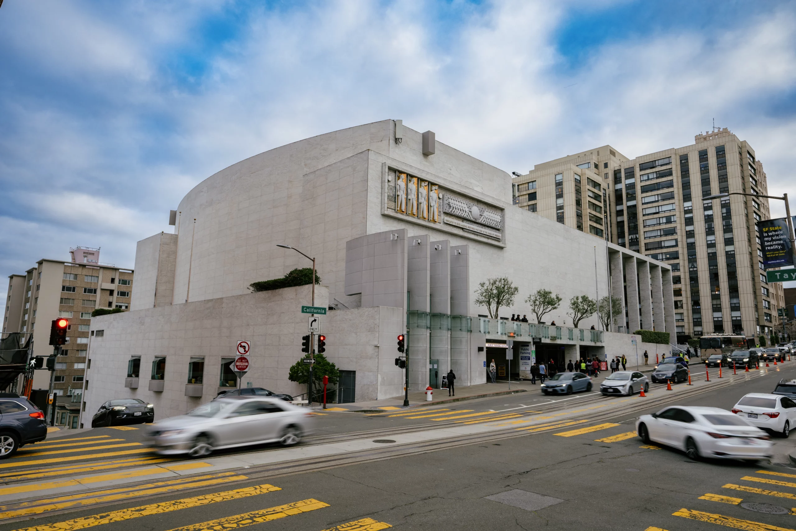 Exterior of large gray modernist building that is the SF Masonic Auditorium, with cars driving along the streets outside in Nob Hill SF