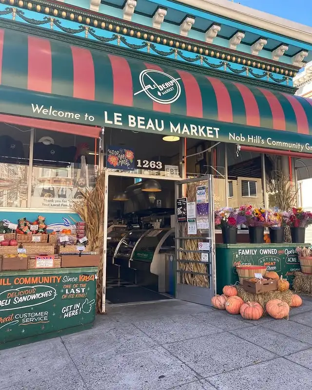 Exterior view of Le Beau Market in Nob Hill in autumn with red and green striped awning that reads ‘Welcome to Le Beau Market’, with pumpkins, fresh flowers, and fruits and veggies for sale out front