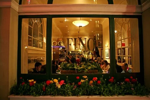 Sidewalk view at night looking into the packed Fino Ristorante dining room, with Fino Ristorante lettering atop the window and a plant box filled with flowers j