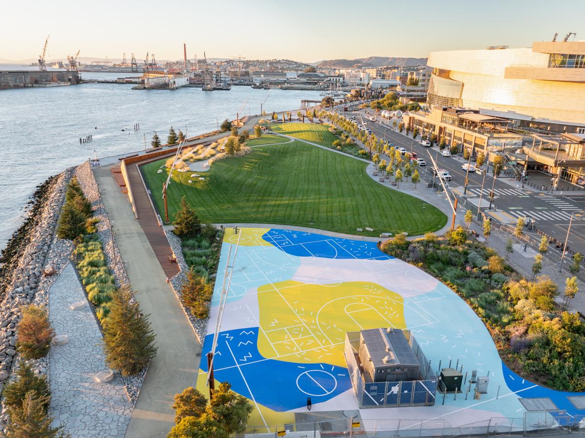 Bird’s eye view of waterfront park with large grassy area, sidewalks, and blue and yellow painted flat cement area