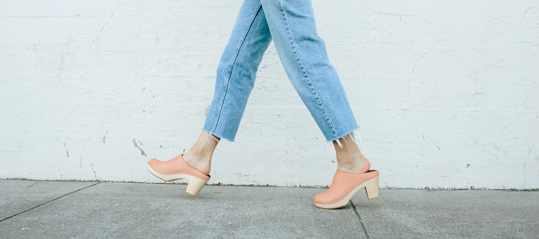 Zoomed in photo showing the lower half of a woman wearing jeans and walking in Bryr Studio’s handcrafted made-to-order tan wooden clogs along a sidewalk in front of a white building wall