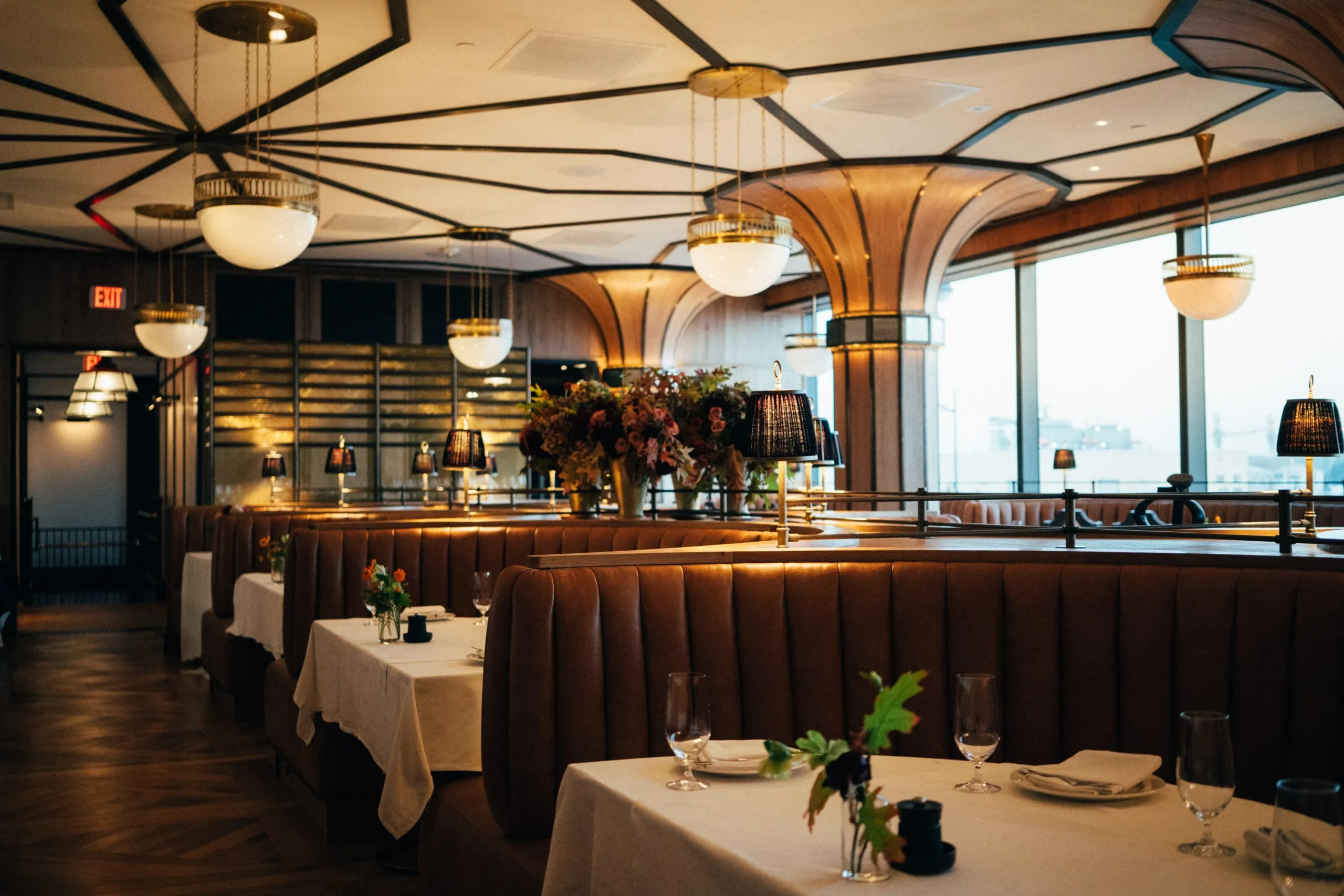 Upscale warmly lit dining room featuring luxe brown leather semi-circle booths, white tablecloths, floor-to-ceiling windows, gold-toned lighting, paneled ceilings, and fresh flowers