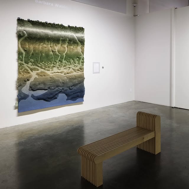 Interior of a gallery space at the Museum of Craft and Design with a handcrafted rug on exhibit, with a contemporary wooden bench for visitors to sit and admire the piece