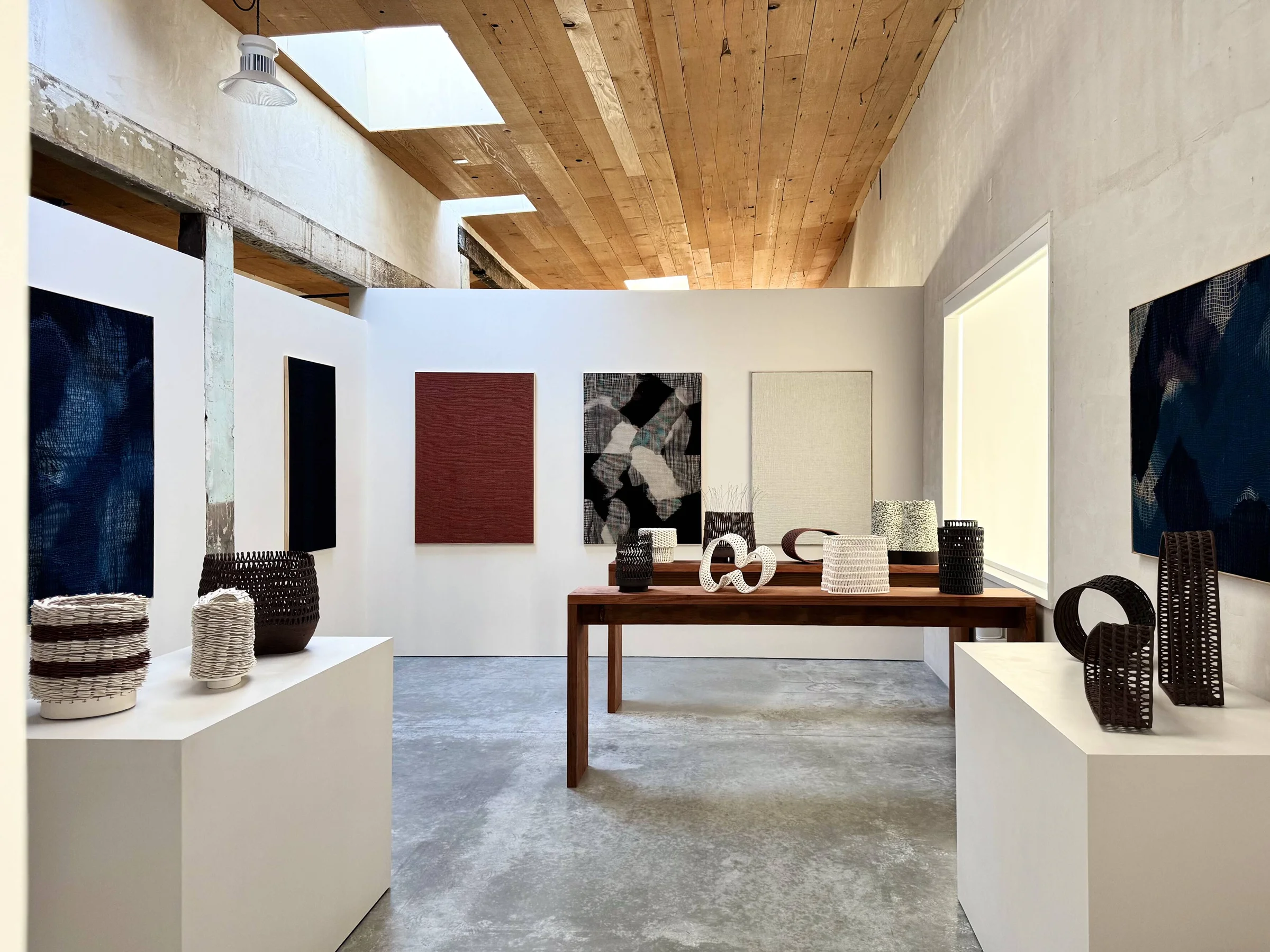 Interior of hugomento store with gray cement floors, soaring wood-paneled ceilings, and displayed modern artwork, baskets, and other artisan household objects displayed on the wall and tall wooden tables or white blocks