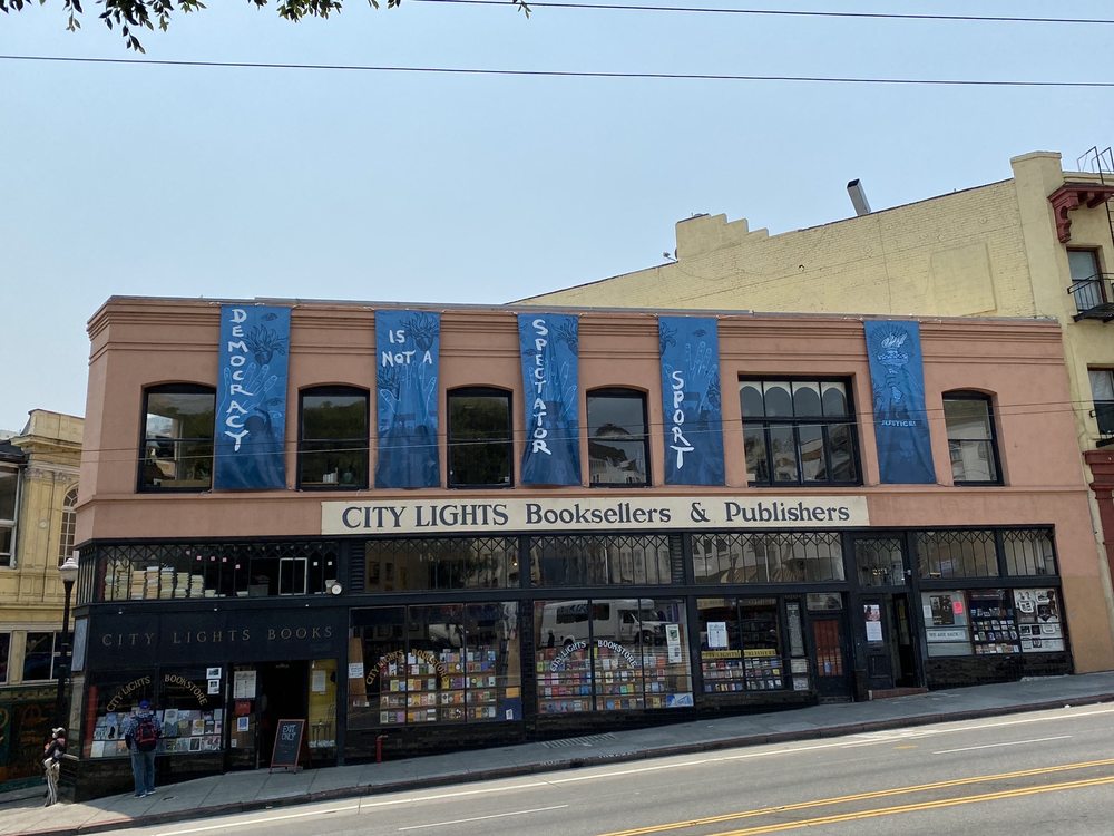 Exterior view from across the street of City Lights Booksellers & Publishers with 5 vertical blue banners across the top wall of building which read: “Democracy Is Not A Spectator Sport”