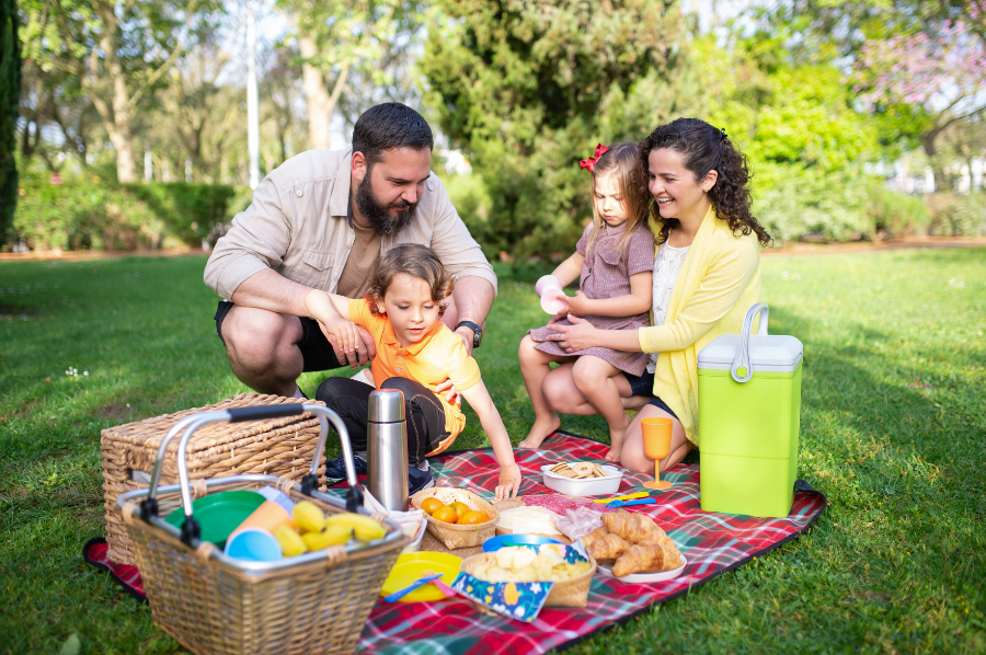 Family enjoying a picnic on a blanket in a sunny park with snacks and a picnic basket.