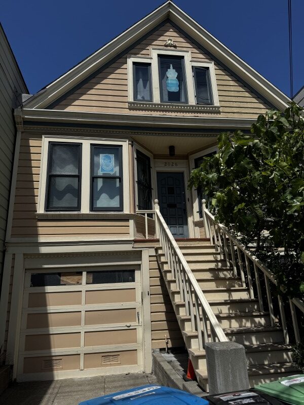 Front of the house before prep, painted tan with cream trim, with decorative metal bars over the lower bay windows and the window beside the front door, a garage door with a row of window panels, and a tree beside the front stairs
