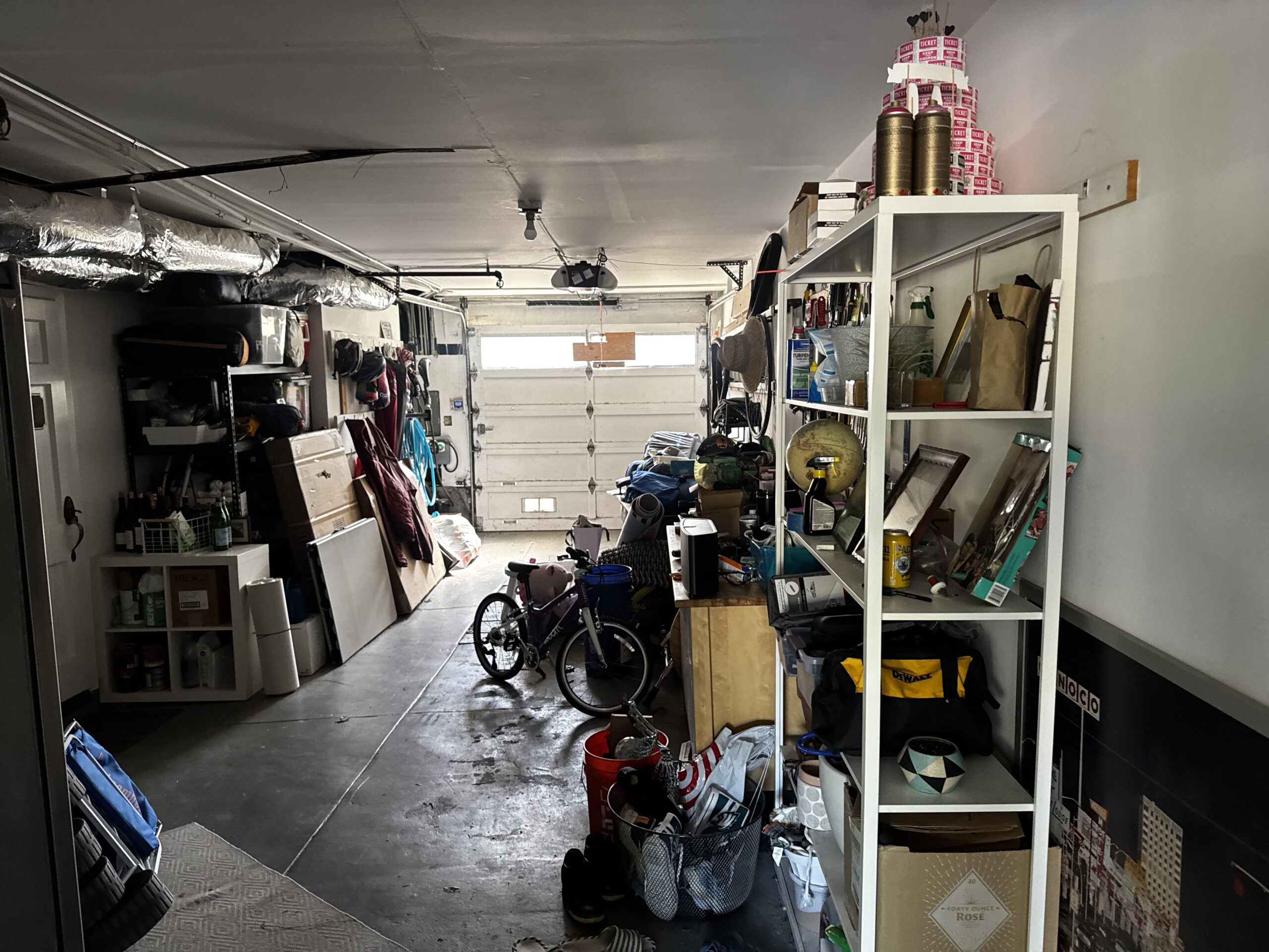 Garage before prep, crowded with shelving, boxes, and a bicycle, looking toward a garage door with a row of window panels