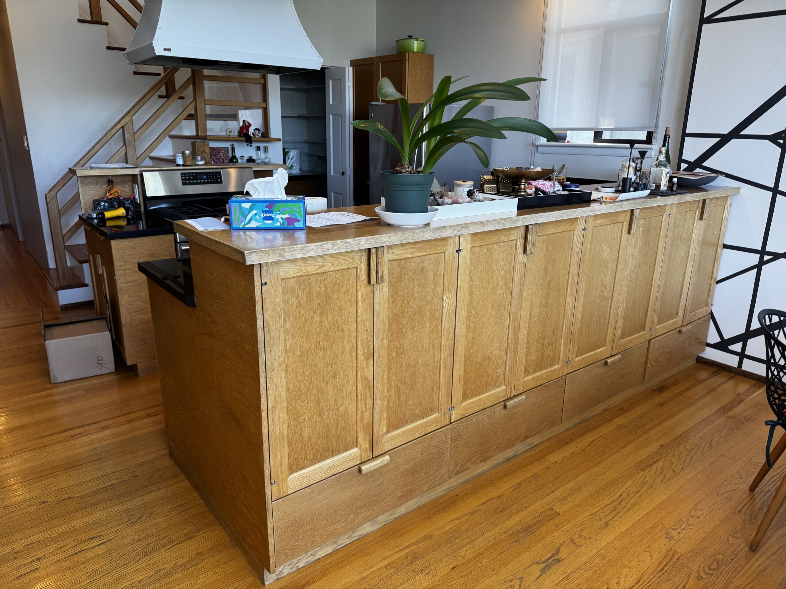 Kitchen peninsula before prep, with natural oak cabinet doors and drawers, a stainless range hood behind it, and an oak stair rail to the left